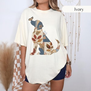 May include: An ivory short-sleeve t-shirt featuring a graphic of a dog in a sitting position. The dog's body is composed of various patchwork patterns in shades of blue, brown, and cream, with a butterfly on its nose and the name Molly printed.