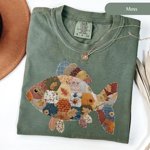 Puede incluir: Una camiseta verde musgo con un diseño de pez patchwork. El pez está hecho de varios patrones de tela en tonos naranja, marrón, azul y amarillo. La camiseta tiene cuello redondo y mangas cortas. También se ve un collar plateado con un pequeño colgante.