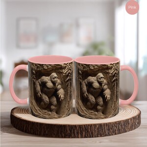 May include: Two pink-handled coffee mugs with a detailed image of a gorilla in a forest setting. The mugs are displayed on a wooden coaster with a natural edge. The interior of the mugs is also pink.