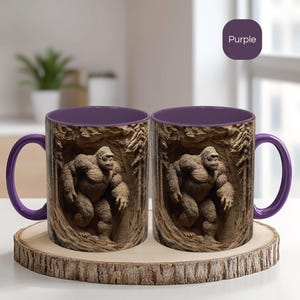 May include: Two ceramic mugs with a purple interior and handle. The exterior features a detailed relief of a Bigfoot figure in a forest setting. The mugs are displayed on a wooden coaster.
