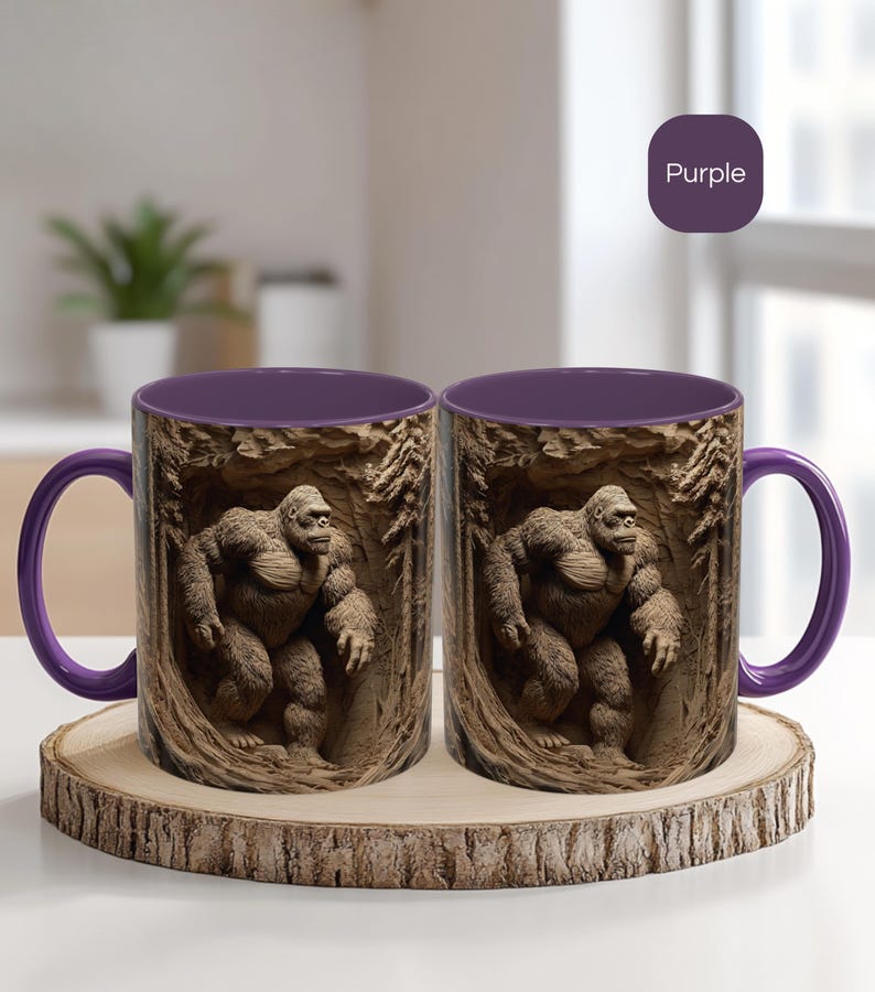 May include: Two ceramic mugs with a purple interior and handle. The exterior features a detailed relief of a Bigfoot figure in a forest setting. The mugs are displayed on a wooden coaster.