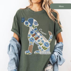 May include: A moss green t-shirt featuring a graphic of a dog silhouette filled with a patchwork pattern of blue flowers, plaid, and solid brown sections. A blue butterfly rests on the dog's nose, and the name Cooper is written in white script.