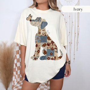 Può includere: Una t-shirt color avorio a maniche corte con una grande grafica di un cane composta da vari riquadri in stile patchwork con motivi floreali e geometrici blu e marroni. Una farfalla poggia sul naso del cane e il nome Molly è stampato in corsivo blu.