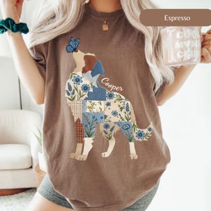 May include: An espresso-colored t-shirt featuring a graphic of a dog composed of blue and white floral and plaid patchwork patterns. A blue butterfly rests on the dog's nose, and the name Cooper is printed in white script above the dog's back.