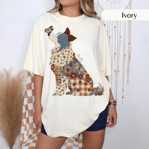 May include: An ivory short-sleeve t-shirt featuring a graphic of a dog in profile. The dog's body is filled with a pattern of autumn leaves, orange flowers, and brown plaid. A butterfly rests on the dog's nose, and the name Molly is printed in script.