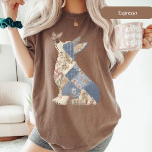 May include: Espresso-colored t-shirt featuring a patchwork dog design with a butterfly on its nose. The dog's body is made of floral and blue patterned fabric. The shirt has a crew neck and short sleeves.