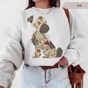 Puede incluir: Una sudadera de cuello redondo color gris ceniza con un gráfico de un caniche compuesto por varios patrones de patchwork de temática otoñal, incluyendo hojas y lunares. Una mariposa descansa sobre la nariz del caniche y el nombre Molly está en azul.