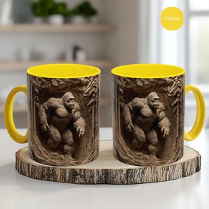 Puede incluir: Dos tazas de cerámica con interior y asa amarillos. El exterior presenta una ilustración detallada de un gorila en un entorno forestal. Las tazas se exhiben sobre un posavasos de madera, con un círculo amarillo que dice "Yellow".