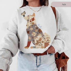 May include: An ash-coloured crewneck sweatshirt featuring a graphic of a dog in a patchwork style with various floral and leaf patterns in shades of brown, tan, and cream. A butterfly rests on the dog's nose, and the name Molly is written in blue.