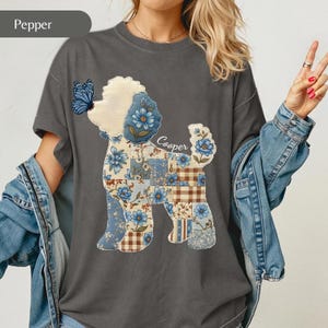 May include: A charcoal grey t-shirt featuring a graphic of a dog silhouette filled with a blue and brown floral and plaid patchwork pattern. A blue butterfly rests on the dog's nose, and the name Cooper is written in white script above the tail.