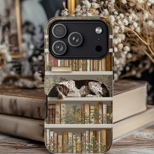 May include: A phone case with a charming design of a dog resting on a bookshelf filled with books and flowers. The case has a clear border and a detailed illustration, creating a cosy, vintage aesthetic. The case is designed for a smartphone.