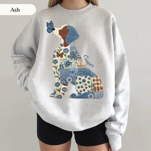 Puede incluir: Una sudadera gris ceniza de cuello redondo con un gráfico grande de un perro compuesto por varios parches de estilo patchwork con motivos florales y cuadros azules y blancos. Una mariposa azul descansa en la nariz y el nombre Cooper está en azul.