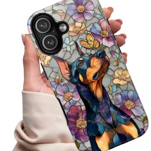 May include: A phone case featuring a stained-glass design of a Doberman Pinscher with a butterfly on its head, surrounded by purple, yellow, and pink flowers. The case has a gray background and a glossy finish.