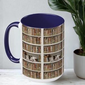 May include: White ceramic mug with a navy blue interior and handle. The mug features a design of a bookshelf with books and two dogs. The dogs are brown and white, and are resting on the shelves.