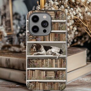 May include: A phone case featuring a bookshelf design with a German Shorthaired Pointer dog resting on books. The case has a white bookshelf with rows of old books and small flowers. The dog is brown and white.