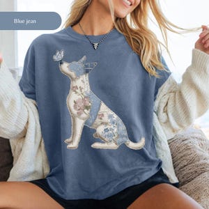 May include: Blue jean crew neck t-shirt with a seated dog design. The dog has floral and blue patterned accents, with a butterfly on its nose. The text "Blue jean" is in a light blue rectangle in the upper left corner.