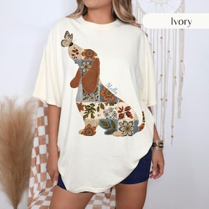 May include: An ivory short-sleeve t-shirt featuring a large graphic of a dachshund silhouette filled with a patchwork pattern of brown, orange, and green floral designs. A butterfly rests on the dog's head, and the name Molly is printed in blue.