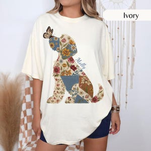 Può includere: Una t-shirt a maniche corte color avorio con la grafica di un cane composto da vari inserti floreali e a fantasia nei toni del blu, marrone e crema. Una farfalla poggia sulla testa del cane e il nome Molly è stampato in corsivo blu.