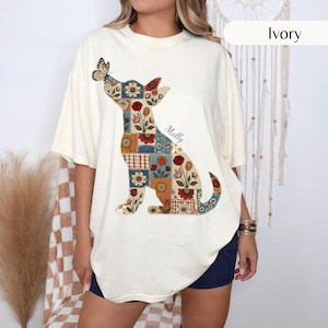 May include: An ivory short-sleeve t-shirt featuring a large graphic of a dog silhouette filled with a colourful floral patchwork pattern. A butterfly rests on the dog's nose, and the name Molly is printed in cursive script next to the dog's back.