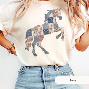 May include: Ivory-colored sweatshirt featuring a patchwork horse design. The horse is composed of various fabric patterns in shades of blue, pink, and cream. The sweatshirt has a crew neck and long sleeves. The word "Ivory" is printed at the bottom.