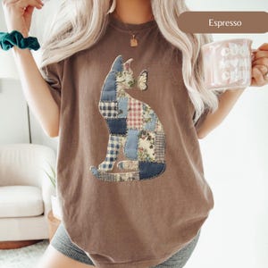 Può includere: T-shirt color espresso con un design a patchwork a forma di gatto e una farfalla. Il gatto è composto da vari modelli di tessuto in stampe blu, rosse e floreali. La maglietta ha una vestibilità comoda.
