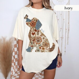 May include: An ivory short-sleeve t-shirt featuring a large graphic of a dog silhouette filled with a patchwork pattern of floral designs, plaid, and a heart. A butterfly rests on the dog's head, and the name Molly is printed in script.