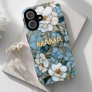 May include: A phone case with a floral design and the word "MAMA" in gold lettering. The case features white flowers, blue and green leaves, and a mosaic-like pattern. The case is designed to fit a smartphone.
