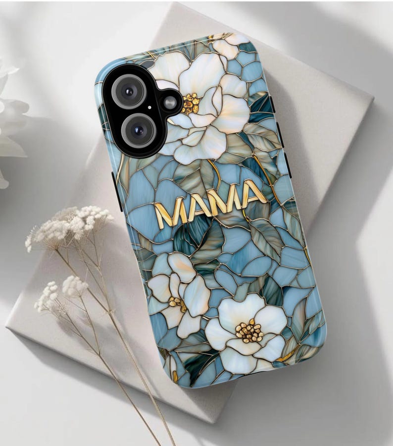 May include: A phone case with a floral design and the word "MAMA" in gold lettering. The case features white flowers, blue and green leaves, and a mosaic-like pattern. The case is designed to fit a smartphone.