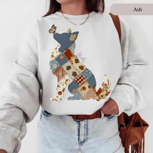 May include: An ash gray crewneck sweatshirt featuring a graphic of a dog in a seated position. The dog's body is filled with a patchwork pattern of blue, orange, and white autumn leaves and plaid squares. A butterfly rests on the dog's nose.