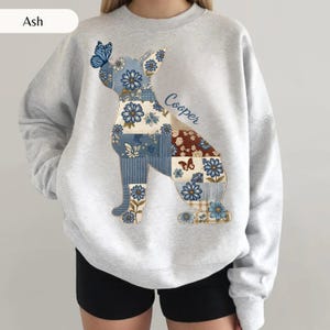 May include: An ash gray crewneck sweatshirt featuring a large graphic of a dog silhouette filled with a blue and brown floral patchwork pattern. A blue butterfly rests on the dog's nose, and the name Cooper is printed in blue script.