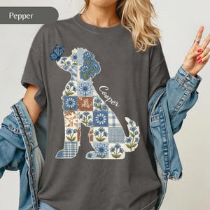 Op de afbeelding: Een antracietgrijs t-shirt met een afbeelding van een hondensilhouet gevuld met een blauw en bruin bloemenpatchworkpatroon. Een blauwe vlinder rust op de neus van de hond en de naam Cooper staat in witte letters naast de hond gedrukt.