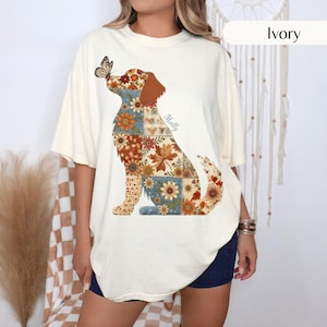 May include: An ivory short-sleeve t-shirt featuring a large graphic of a dog composed of various floral and leaf patterns in shades of orange, brown, and blue. A butterfly rests on the dog's nose, and the name Molly is printed in script.