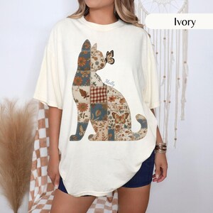 Puede incluir: Una camiseta de manga corta color marfil con un gráfico grande de un gato sentado compuesto por un patrón de patchwork. El diseño del gato incluye motivos florales, de hojas y de cuadros en tonos tierra, con una mariposa cerca de la nariz y el nombre Molly.