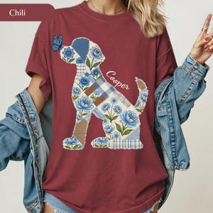 May include: A chili red t-shirt featuring a graphic of a dog silhouette filled with a blue floral and plaid patchwork pattern. A small blue butterfly rests on the dog's nose, and the name Cooper is printed in white script next to the dog's back.