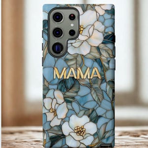 May include: A phone case featuring a floral design with white flowers and green leaves on a blue background. The word "MAMA" is displayed in gold lettering. The case has a glossy finish.