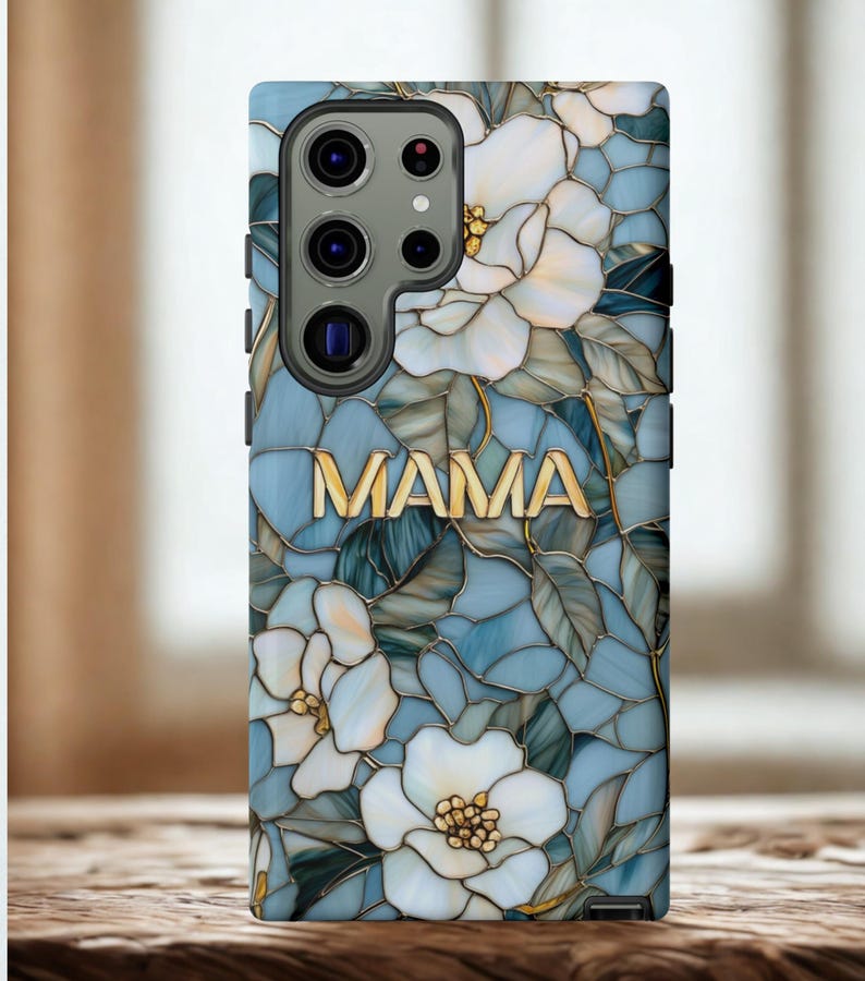 May include: A phone case featuring a floral design with white flowers and green leaves on a blue background. The word "MAMA" is displayed in gold lettering. The case has a glossy finish.