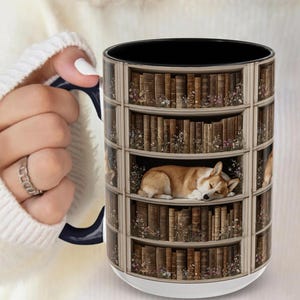 May include: A white ceramic mug with a black interior and handle. The mug features a design of a bookshelf with old books and a sleeping corgi dog. The mug is held by a person with a white sweater and a silver ring.