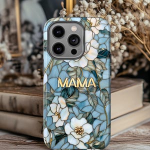 May include: A phone case with a floral design and the word "MAMA" in gold lettering. The case features white flowers, blue and green leaves, and a stained-glass effect. The case is designed for a smartphone.