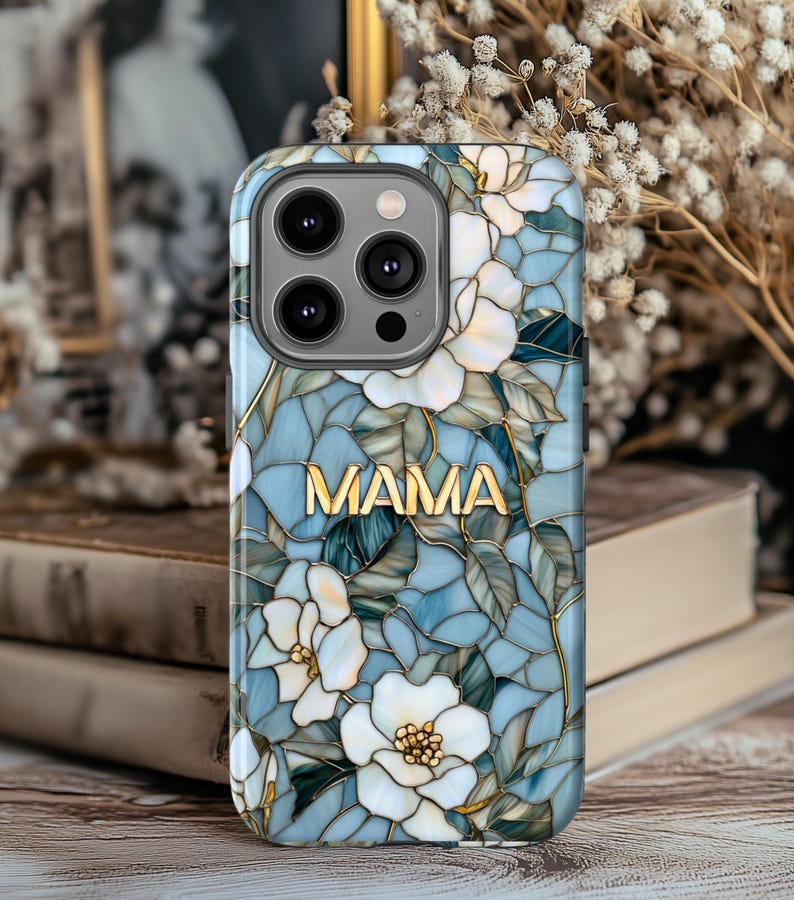 May include: A phone case with a floral design and the word "MAMA" in gold lettering. The case features white flowers, blue and green leaves, and a stained-glass effect. The case is designed for a smartphone.
