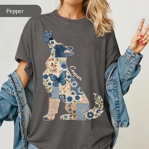 May include: A charcoal gray t-shirt featuring a graphic of a dog silhouette filled with a patchwork pattern of blue flowers, plaid squares, and butterflies. The name Cooper is printed in white script next to the dog. The shirt has short sleeves.