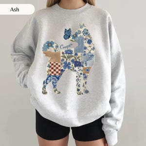 May include: An ash gray crewneck sweatshirt featuring a large graphic of a dog silhouette filled with a blue and white floral patchwork pattern. A blue butterfly sits on the dog's head, and the name Cooper is written in blue script text.