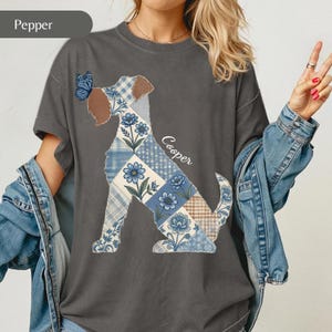 May include: A charcoal grey t-shirt featuring a graphic of a dog silhouette filled with a blue and white patchwork pattern of flowers and plaid. A blue butterfly rests on the dog's head, and the name Cooper is printed in white script.
