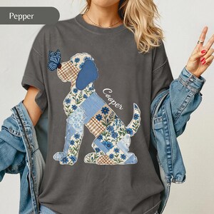 May include: A charcoal grey crewneck t-shirt featuring a graphic of a dog in a patchwork style with blue floral, plaid, and solid blue patterns. A blue butterfly rests on the dog's nose, and the name Cooper is printed in white script.