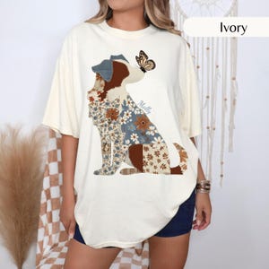 Può includere: Una t-shirt color avorio con la grafica di un cane composto da vari inserti con motivi floreali e di foglie nelle tonalità del marrone, blu e bianco. Una farfalla poggia sul naso del cane e il nome Molly è stampato in corsivo.