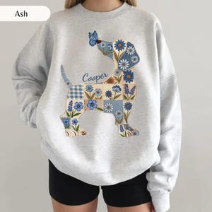 May include: An ash grey crewneck sweatshirt featuring a graphic of a dachshund silhouette filled with a patchwork pattern of blue flowers, checkered squares, and a blue butterfly on its nose. The name Cooper is printed in blue script above the dog.