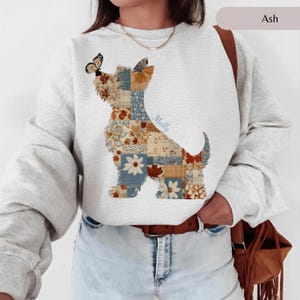 Peut inclure: Un sweat-shirt à col rond gris cendré orné d'un grand motif représentant la silhouette d'un chien remplie d'un patchwork de motifs floraux et à carreaux bleus, orange et blancs. Un papillon est posé sur la tête et le prénom Molly est inscrit.