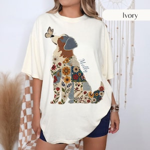 May include: An ivory t-shirt featuring a graphic of a dog with a patchwork-style body filled with floral patterns and a butterfly perched on its nose. The name Molly is printed in blue script next to the dog. The shirt has short sleeves.