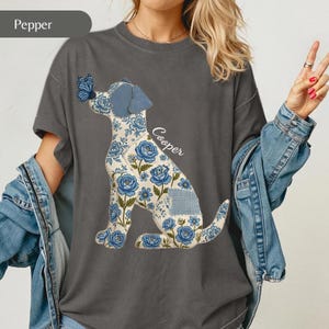May include: A charcoal grey t-shirt featuring a graphic of a dog in a sitting position. The dog has a blue floral pattern and a blue butterfly on its nose. The name Cooper is printed in white script next to the dog. The shirt has a crew neckline.