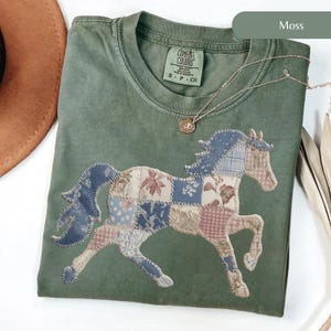 May include: A moss green t-shirt featuring a patchwork horse design. The horse is made of various fabric patterns in blue, pink, and cream. A gold necklace with a pendant rests on the shirt. The shirt has a Comfort Colors label.