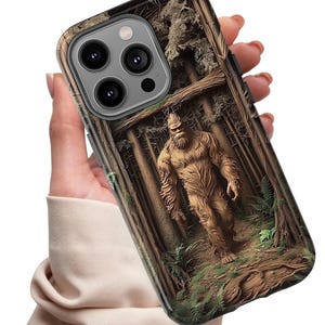 May include: A phone case featuring a detailed illustration of Bigfoot in a forest setting. The design showcases a brown Bigfoot figure amidst tall trees and foliage, with a textured, wood-like appearance. The case has a dark border and camera cutouts.